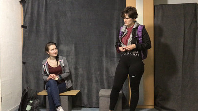 The two actors talk to each other, one sitting, one standing. 