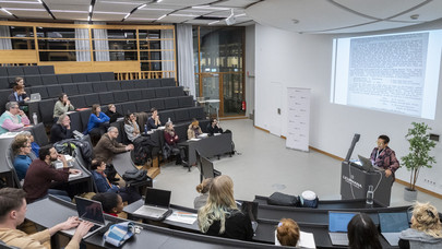 A photograph of a small lecture hall, at the speakers desk one can see Yvette Christiansë looking at her script. There are 24 people sitting in the seats that form a ascending half circle around the room 