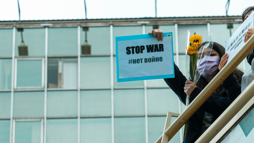 Mädchen hält Plakat mit der Aufschrift “Stop War” von einer Brüstung. In der anderen Hand hält sie einen Strauß mit Sonnenblumen.