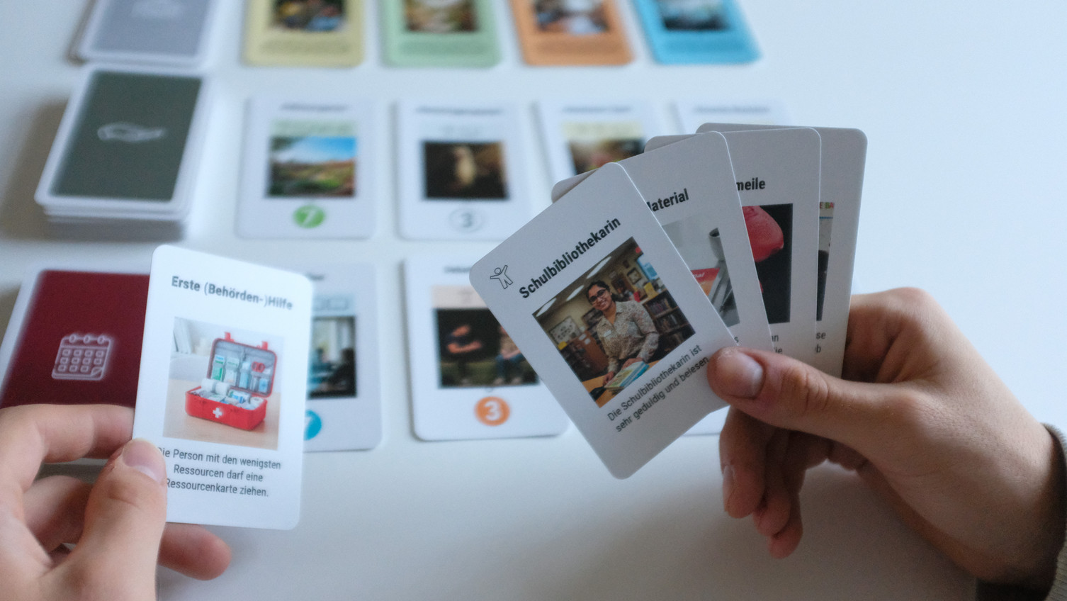 Two hands holding playing cards. Behind them, more playing cards are spread out on a table.
