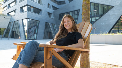 Paula Muche, Studierende des Masters Medien und Digitale Kulturen, sitzend auf einem Holzstuhl vor dem Zentralgebäude der Leuphana.