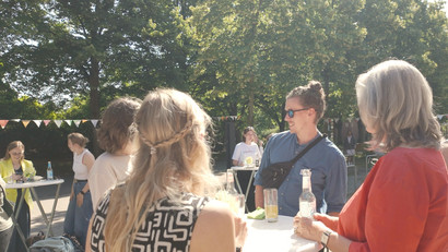 Ein Foto von den Graduate School Homecoming Days 2025 vor Gebäude 14 auf dem Campus der Leuphana: Vier Personen bei sommerlichem Wetter im Gespräch an einem Stehtisch, auf dem Getränke stehen. 