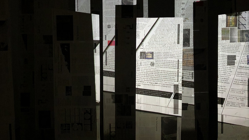 you see printed paper webs spread out as sculptures in the room, on them a projection of a hand leafing through the newspaper.