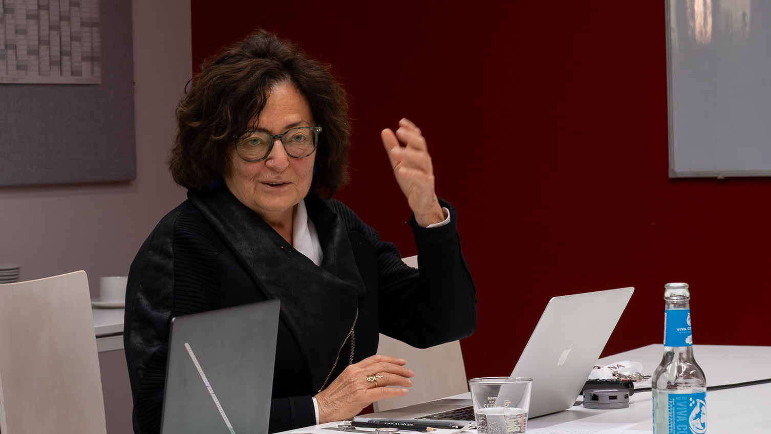 Workshop Koch Eine Frau spricht an einem Tisch sitzend und hebt dabei die linke Hand