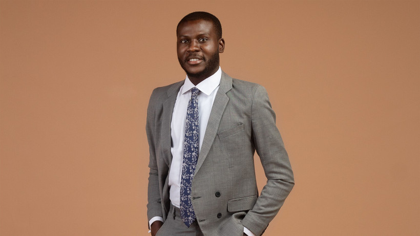 Portrait of ILGSPD-Student Damilola Michael Oguntade in a gray suit in front of a reddish brown background.