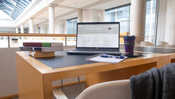 Ein Tischarbeitsplatz, auf dem ein Laptop steht. Davor ein Stuhl, über dem ein Pullover hängt. Im Hintergrund sieht man das übrige Foyer mit Säulen.
