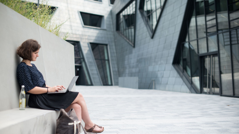 A student with a laptop on her lap working in front of the central building of Leuphana University.