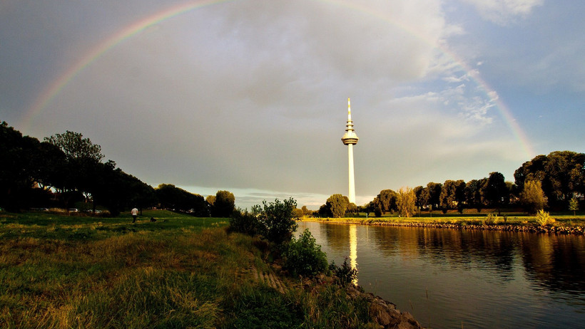 Fluss mit Fernsehturm im Hintergrund und einem Regenbogen vor Wolkenverhangenen Himmel