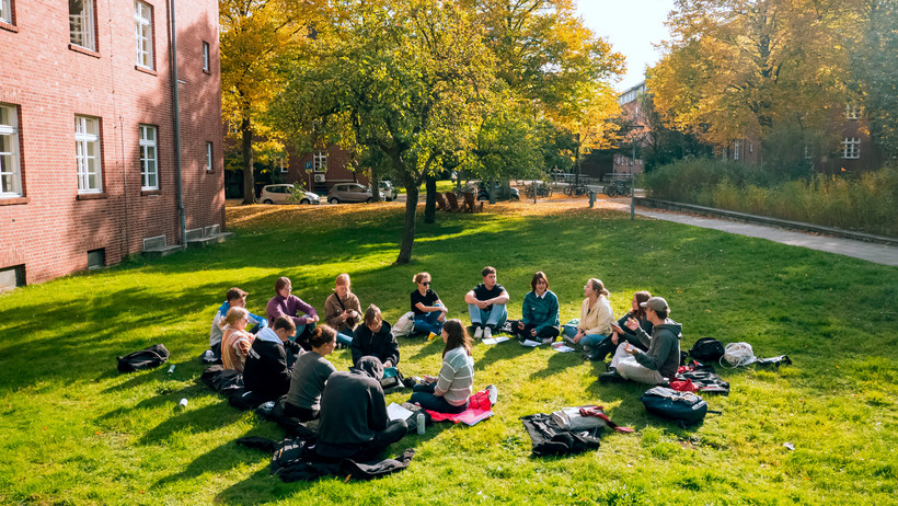 Eine Gruppe Studierender sitzt bei Sonnenschein draußen im Gras