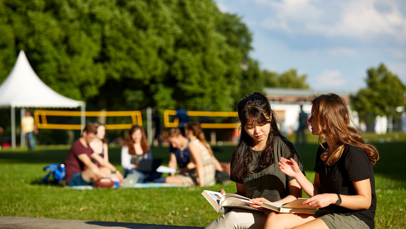 Zwei Studierende sitzen draußen auf dem Campus und schauen in ein Fachbuch.