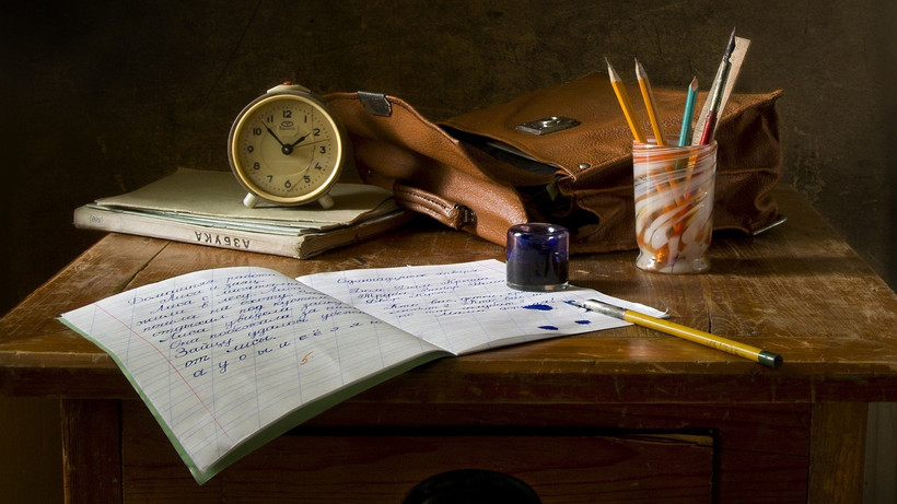 Ein Heft auf einem antiken Schultisch. Im Hintergrund eine braune Ledertasche, ein Wecker und ein Glas, in dem Stifte stehen.