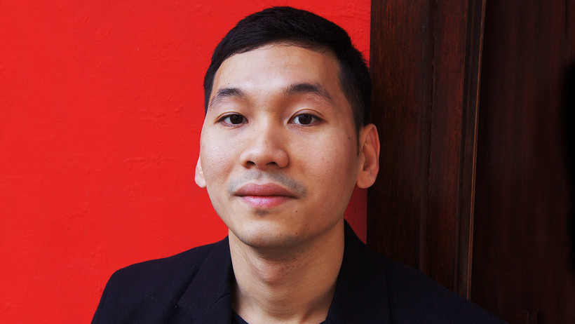 [Translate to Englisch:] Tuan Nghuyen stands in front of a red wall in a black jumper.