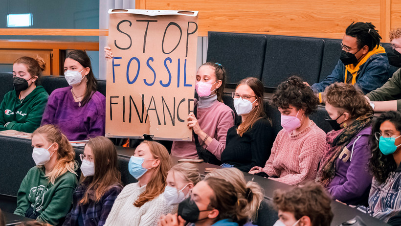 Eine Studierende hält bei einer Podiumsdiskussion ein Plakat gegen Fossil Fuels hoch