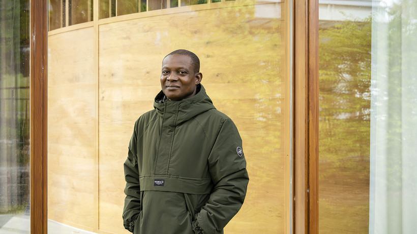 LIAS Fellow Idowu Emmanuel Adeniyi in grüner Jacke vor einer Glaswand, durch die man in einen Hörsaal der Universität gucken kann