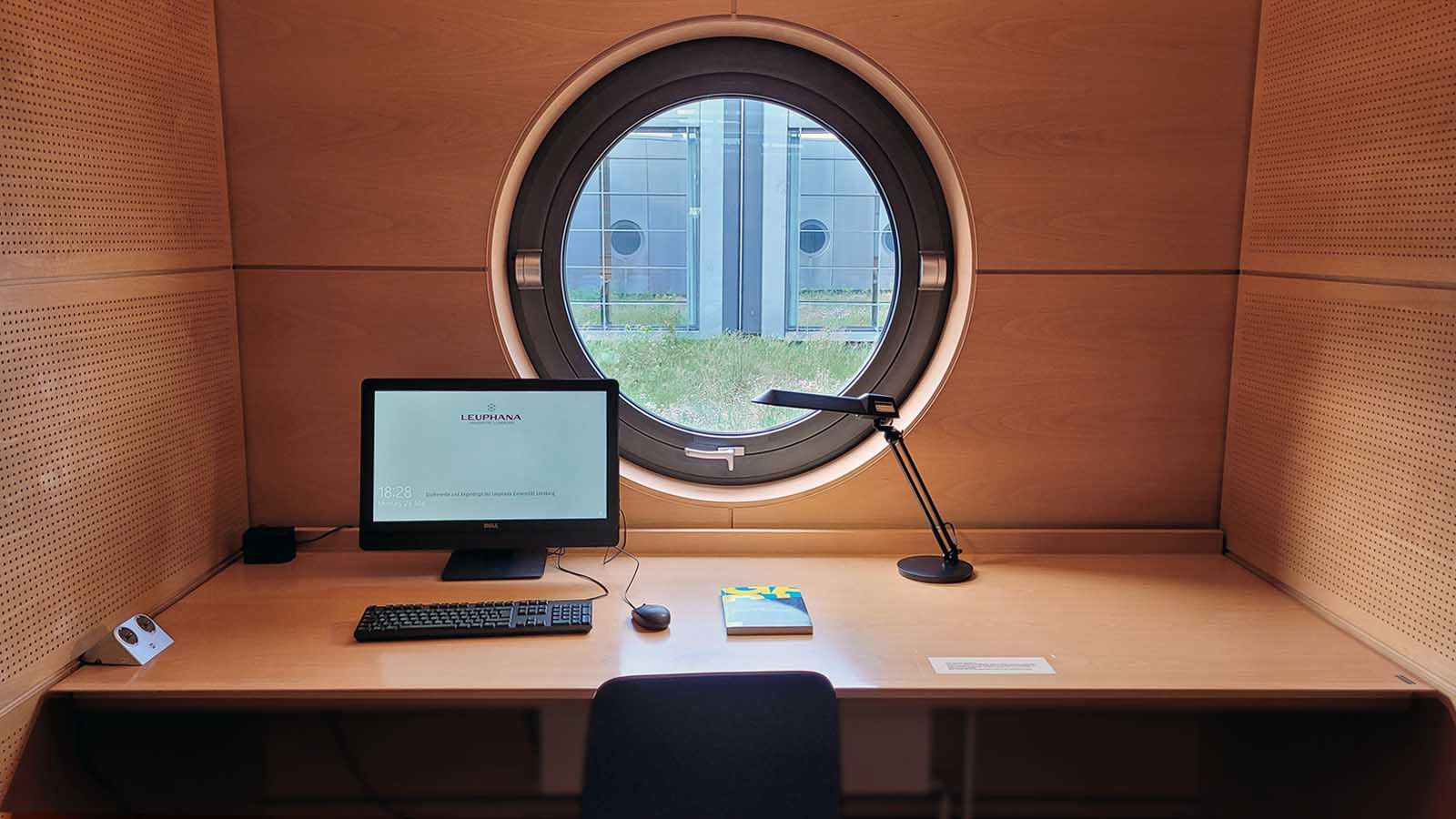 Einzelarbeitsraum in der Bibliothek mit Schreibtisch, Schreibtischlampe und PC. Im Hintergrund des von innen holzvertäfelten Raumes ein modernes Rundfenster mit Blick auf das begrünte Dach